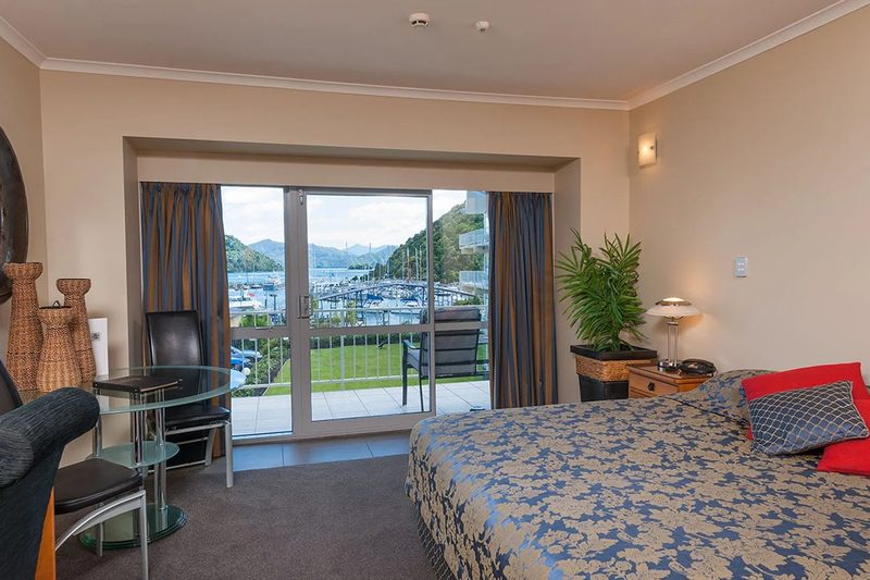 Guest room with a queen bed, dining table, patio doors and a view across Picton marina.