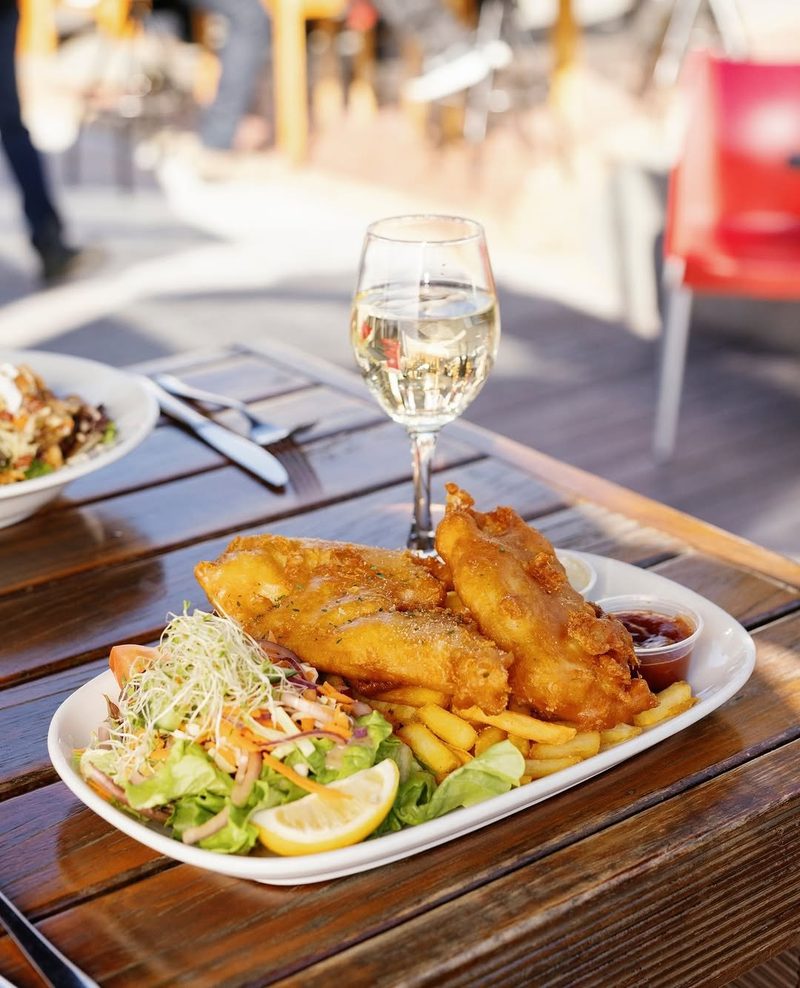 Large plate of battered fish and chips with salad, lemon, sauces and a glass of white wine.