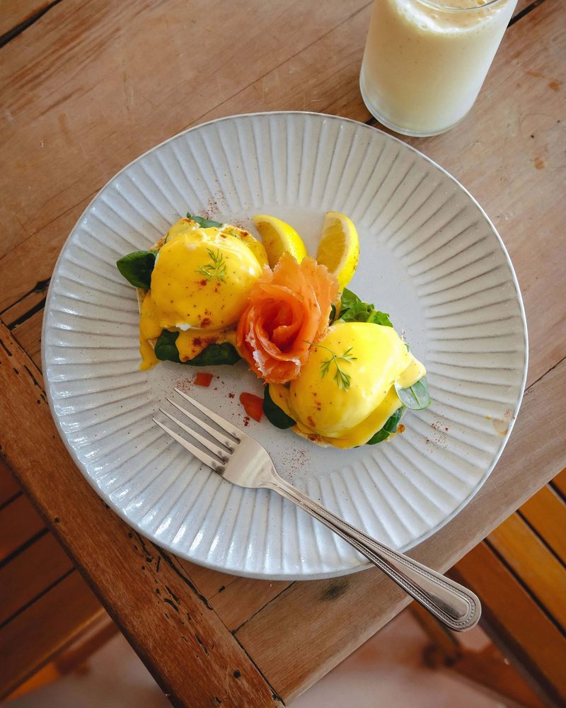 Eggs Benedict with smoked salmon, spinach, hollandaise, lemon, and a smoothie on a wooden café table.