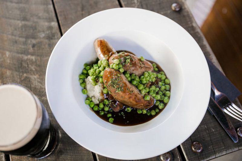 Bangers and mash with peas and onion gravy, photographed on a rustic table beside a dark beer.