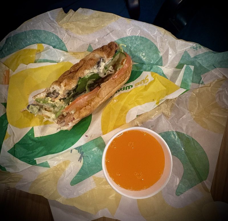 A Subway sandwich with salad fillings and an orange drink on branded wrapping.