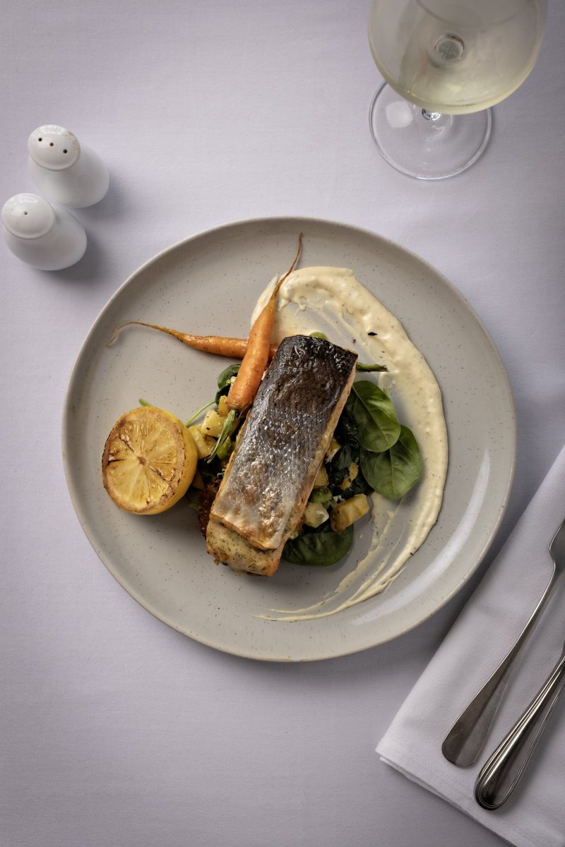 Plated salmon with roasted carrots, greens, lemon and sauce on a white-clothed table.
